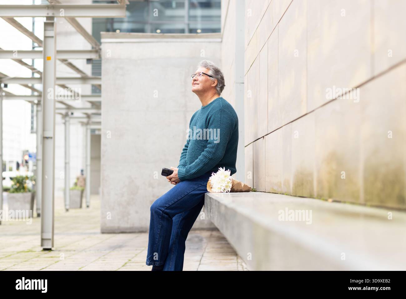 Senior-Mann sitzt auf einer Betonbank in der plaza und hält ein schwarzes Gerät mit weißem Blumenstrauß, Kopierraum Stockfoto