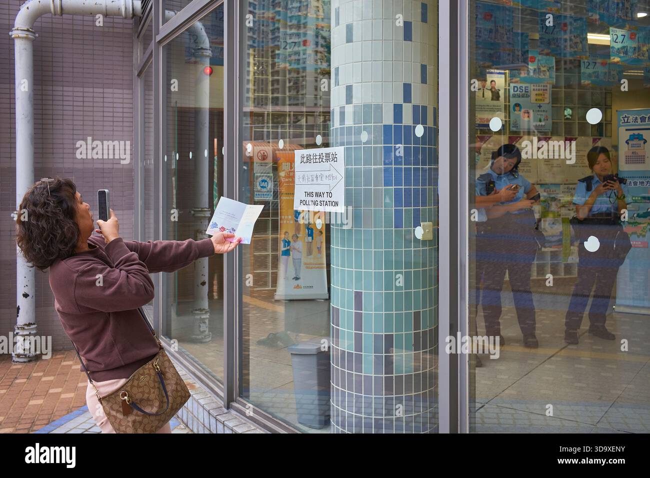 Hongkong, China. Dezember 2025. Eine Wählerin fotografiert ihre Wahlurkunde vor einem Wahllokal und Polizeibeamte überwachen den reibungslosen Ablauf der Wahlen in Tin Shui Wai. Die Parlamentswahlen 2025 in Hongkong finden am 7. Dezember 2025 statt, um die 90 Mitglieder des 8. Hongkonger Legislativrates zu wählen. Quelle: SOPA Images Limited/Alamy Live News Stockfoto