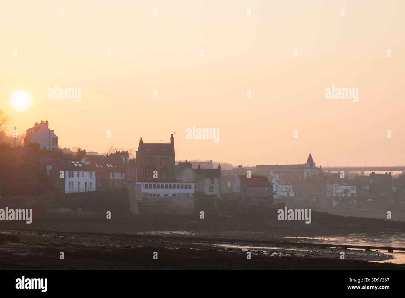 Goldenes Sonnenuntergangslicht taucht die historischen Gebäude an der Küste von South Queensferry neben dem Firth of Forth auf, mit einem trüben Blick auf die Forth Road Brid Stockfoto