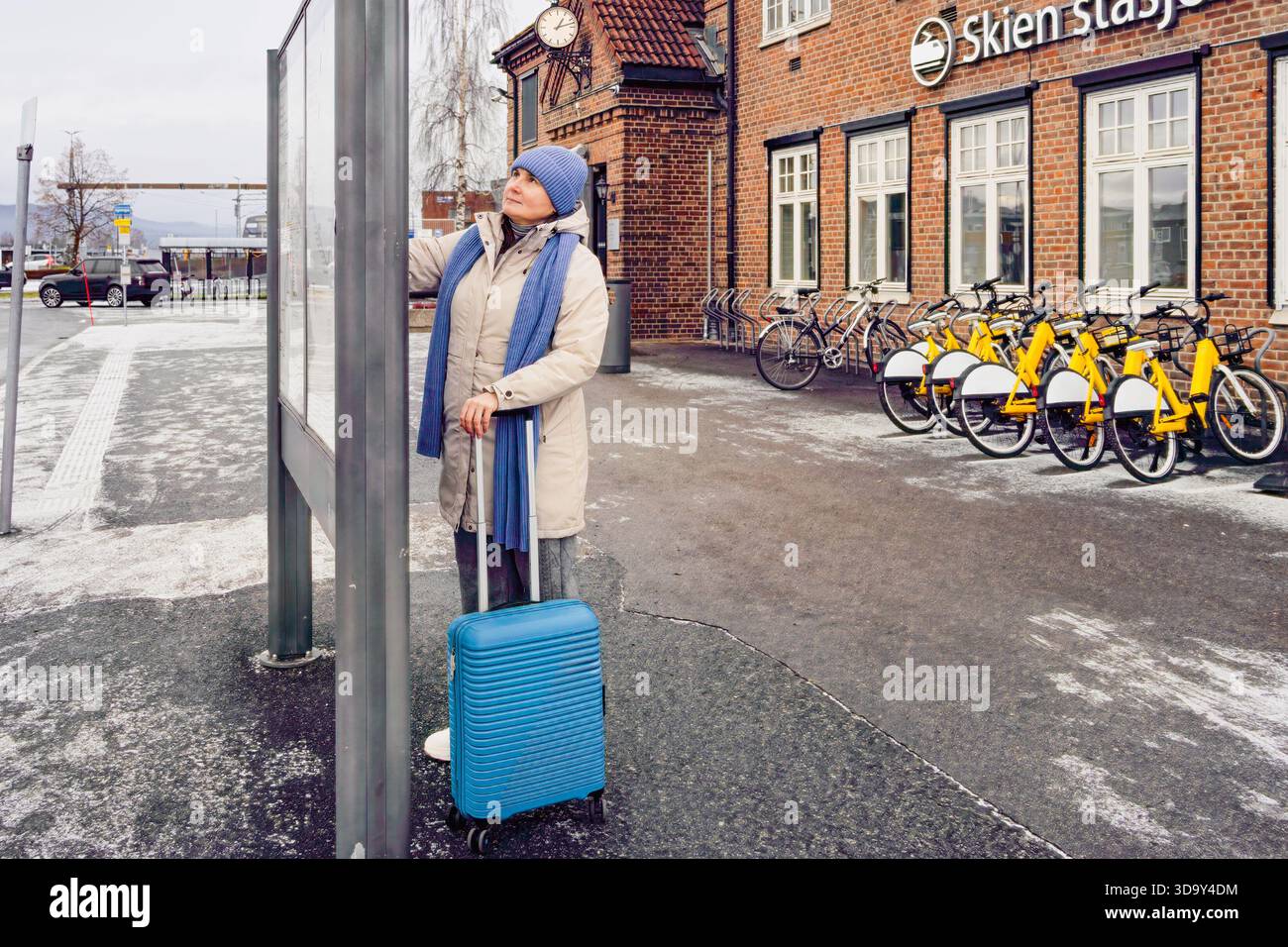 Reisende mittlerer Alters mit blauem Gepäck schauen sich an der Haltestelle den Busfahrplan an, warm gekleidet in Winterkleidung, mit Fahrrädern und einer Backsteinstruktur Stockfoto