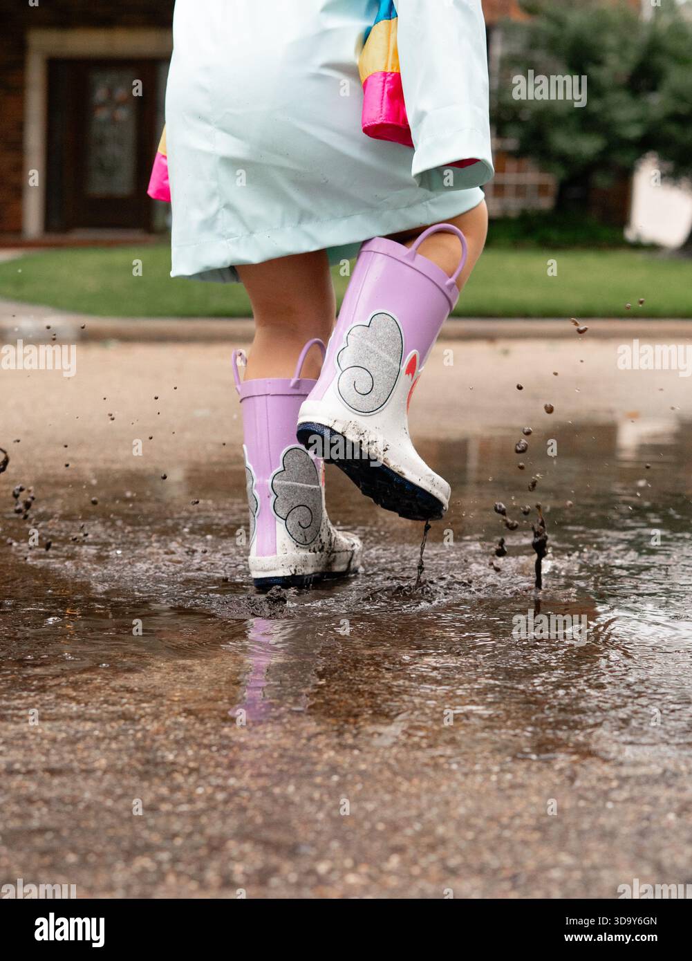 Kind, das in einer schlammigen Pfütze mit bunten Einhorn-Regenstiefeln planscht, während es auf einer nassen Straße springt Stockfoto
