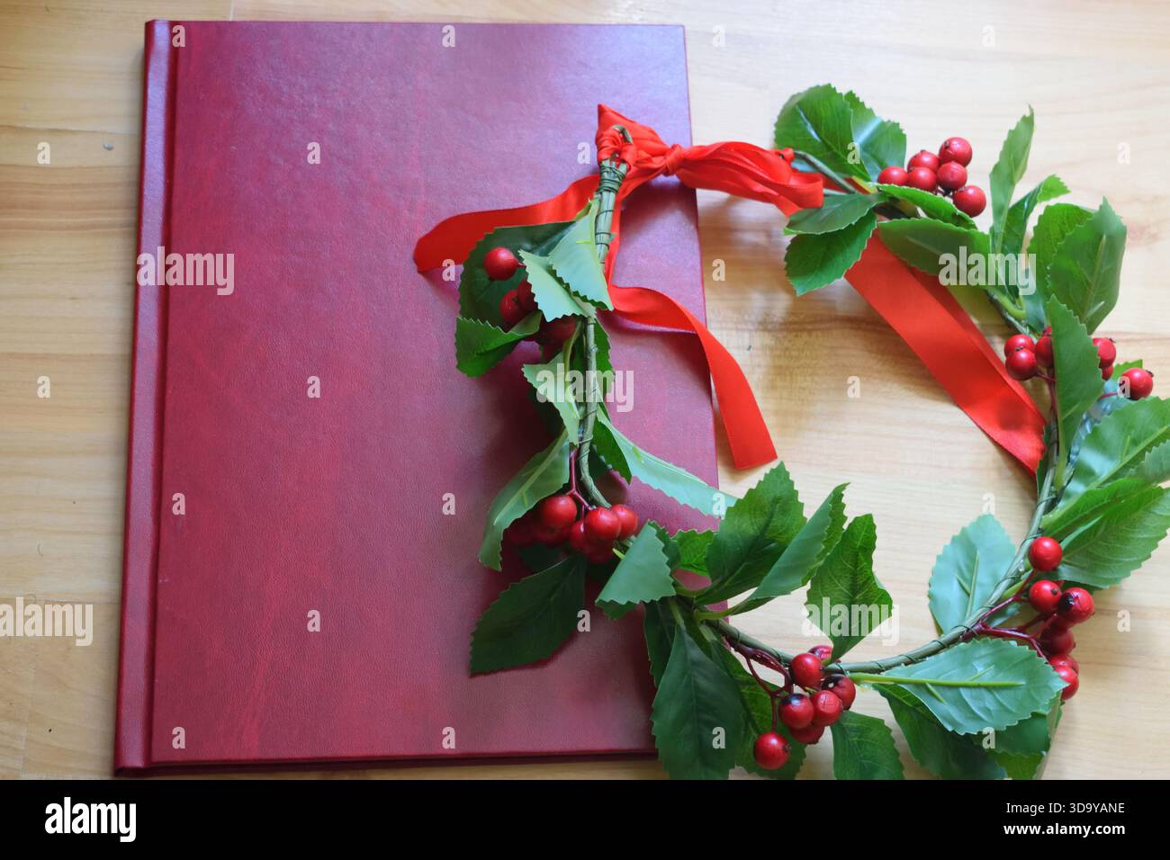Rotes Abschlussbuch auf einem Holztisch neben einem Lorbeerkranz mit grünen Blättern, roten Beeren und einem roten Band, das ein akademisches d symbolisiert Stockfoto