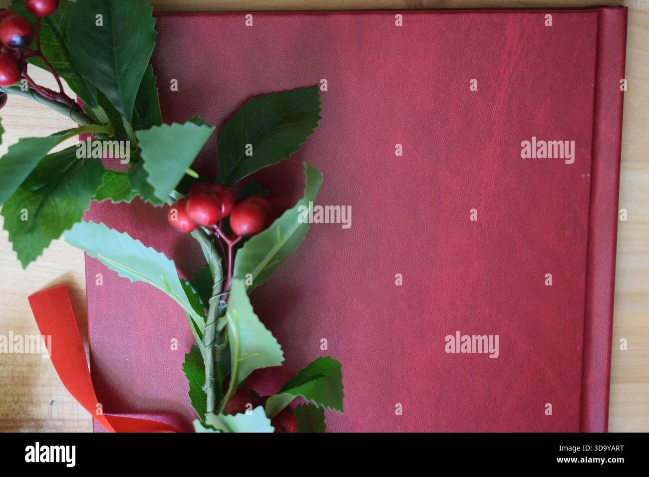 Rotes Abschlussbuch auf einem Holztisch neben einem Lorbeerkranz mit grünen Blättern, roten Beeren und einem roten Band, das ein akademisches d symbolisiert Stockfoto