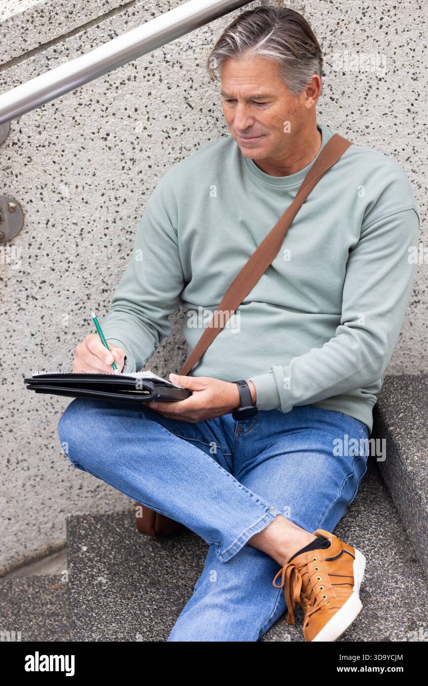 Senior Mann schreibt in Notizbuch mit grünem Stift auf Außentreppen am Handlauf mit Kuriertasche Stockfoto