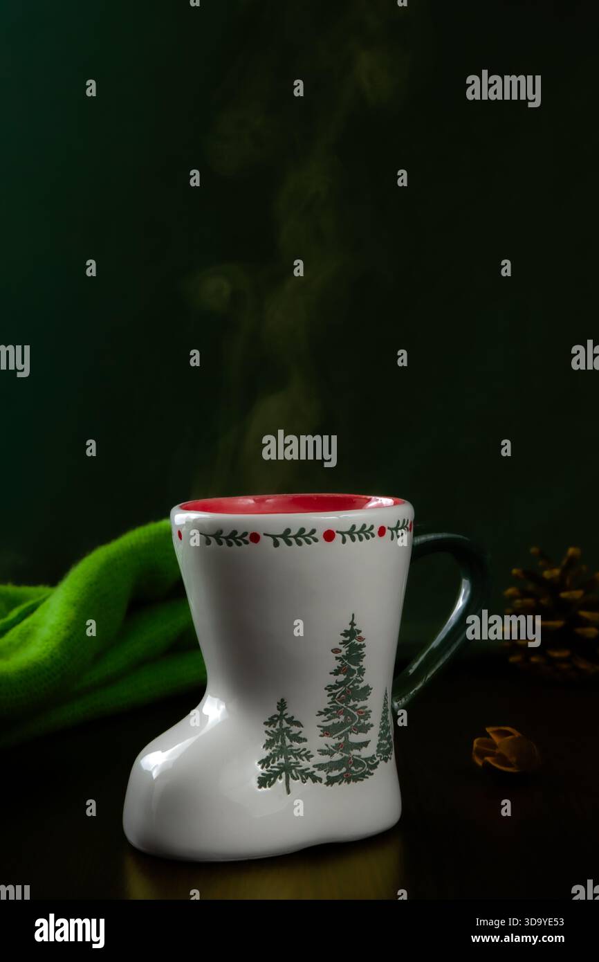 Weißer Keramikbecher in Form eines Stiefels, mit Abbildungen von grünen Tannenbäumen und einem dekorativen stechpalmenmuster mit roten Beeren am Rand. Stockfoto