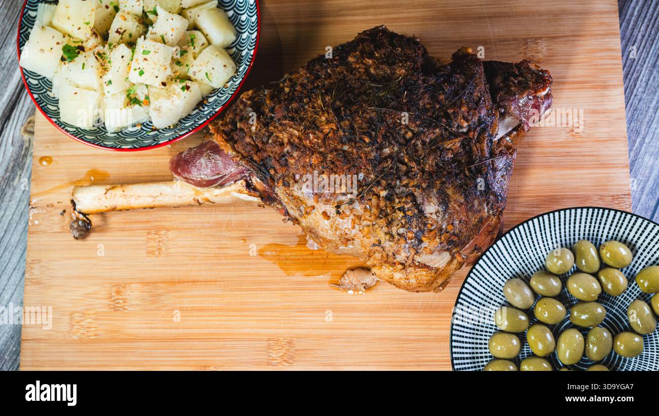 Gebratene Lammkeule auf einem Brett mit Beilagen: Oliven und Maniok-Pommes. Golden, gewürzt, perfekt für ein komplettes und herzhaftes festliches Essen. Stockfoto