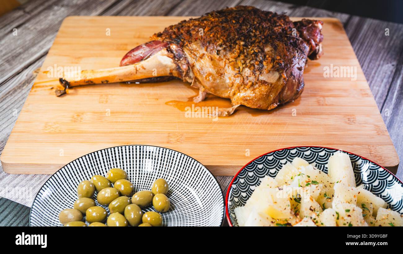 Gebratene Lammkeule auf einem Brett mit Beilagen: Oliven und Maniok-Pommes. Golden, gewürzt, perfekt für ein komplettes und herzhaftes festliches Essen. Stockfoto