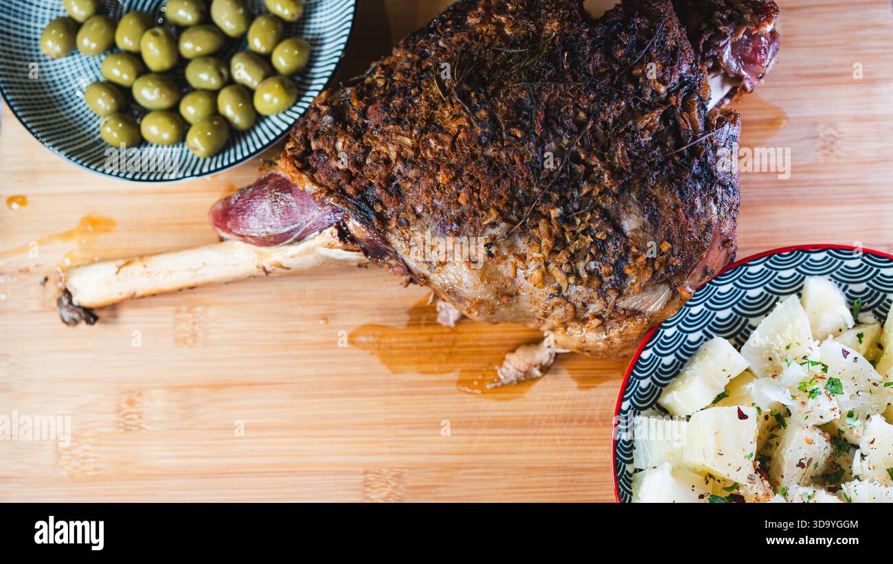 Gebratene Lammkeule auf einem Brett mit Beilagen: Oliven und Maniok-Pommes. Golden, gewürzt, perfekt für ein komplettes und herzhaftes festliches Essen. Stockfoto