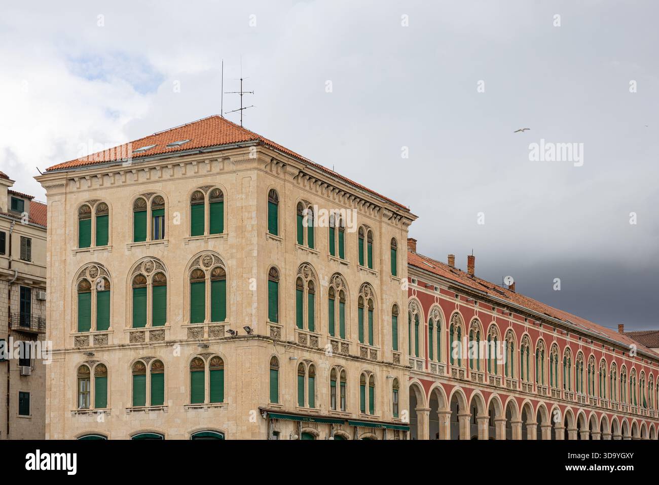 Ein großes, sonnendurchflutetes historisches Gebäude mit Bogenfenstern, grünen Fensterläden und einer langen Arkade, die zeitlosen Charme und mediterrane Architektur vermittelt Stockfoto