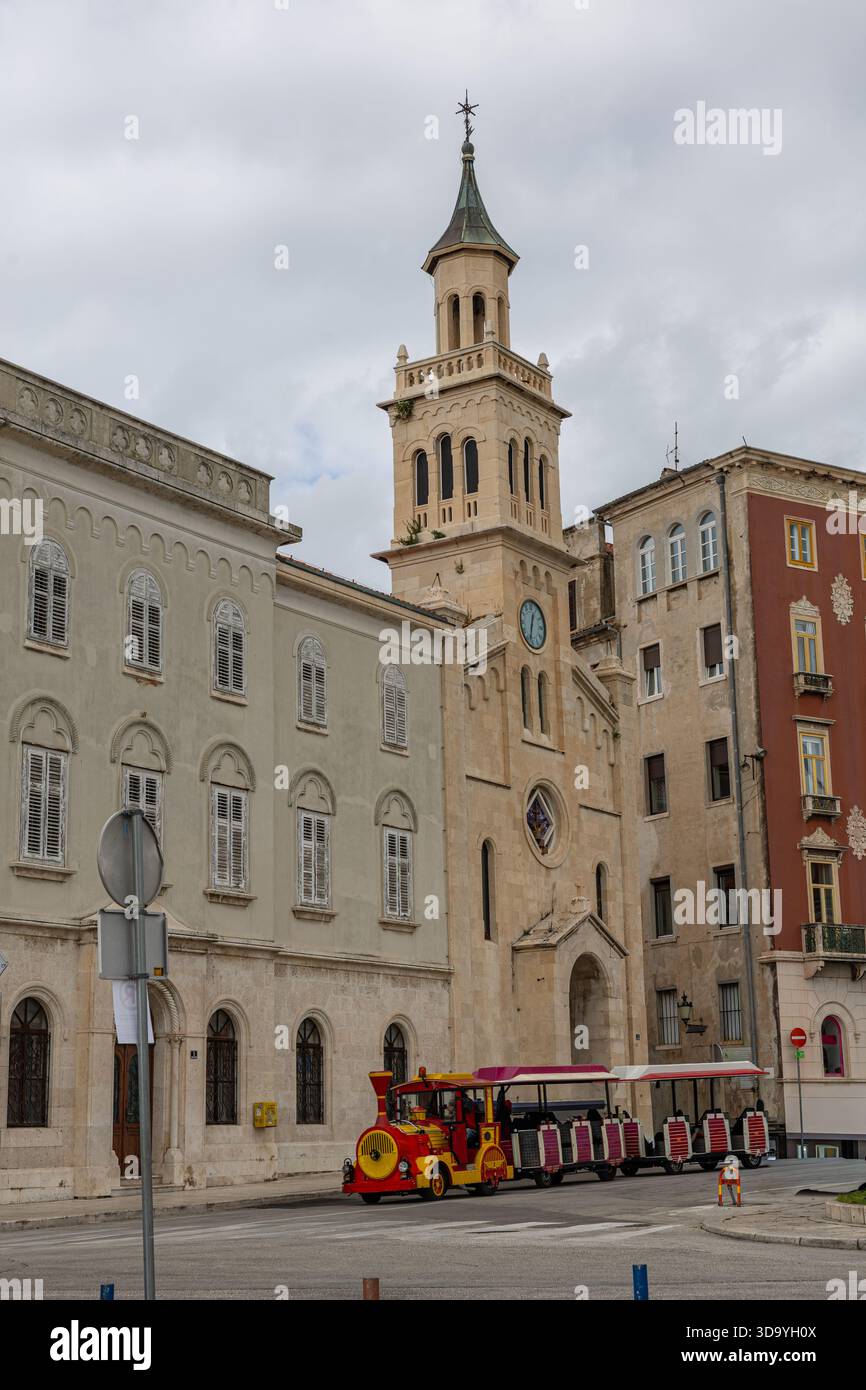 Eine historische europäische Straßenszene mit einer beigefarbenen Steinkirche mit einem hohen Uhrenturm, flankiert von alten Gebäuden. Ein leuchtend roter Touristenzug sitzt Stockfoto