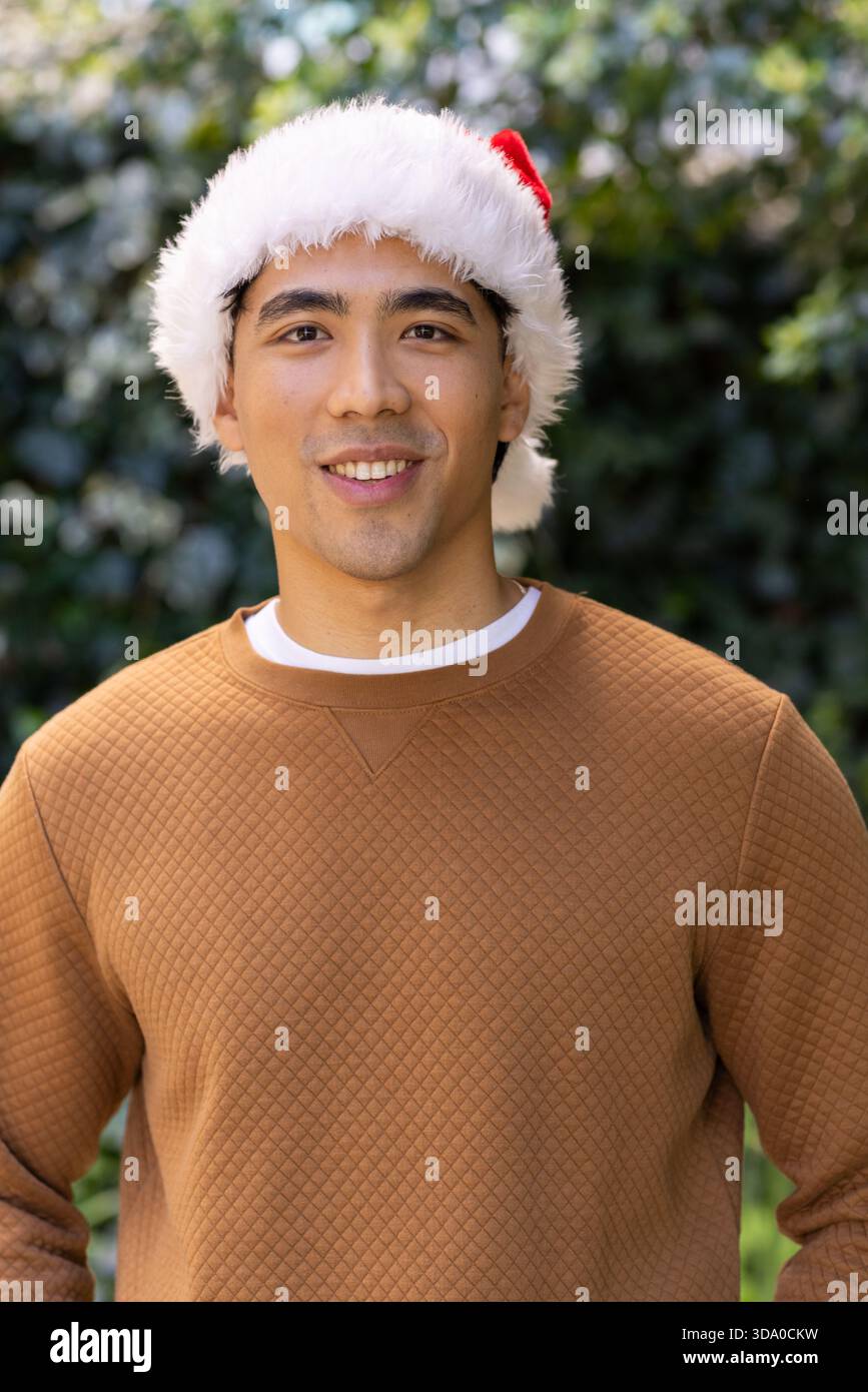 Asiatischer Mann an grüner Hecke mit rotem Weihnachtsmann-Hut, braunem gestepptem Pullover und weißem T-Shirt Stockfoto