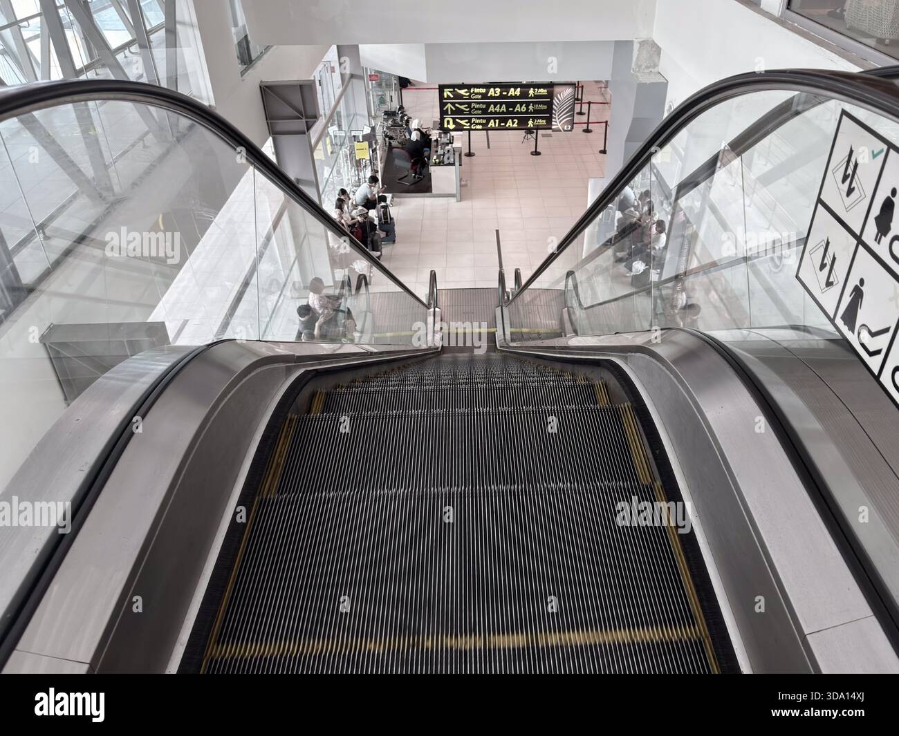 Penang, Malaysia - 28. April 2025: Rolltreppe führt hinunter in den Terminalbereich mit sichtbarer Gate-Anzeige für A1-A6. Reisende werden an den Check-in-Schaltern gesehen Stockfoto