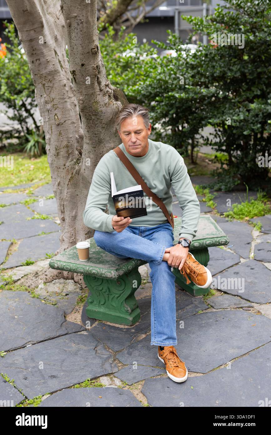 Reifes männliches Lesebuch auf der Hofbank mit Kuriertasche und Einweg-Kaffeetasse Stockfoto