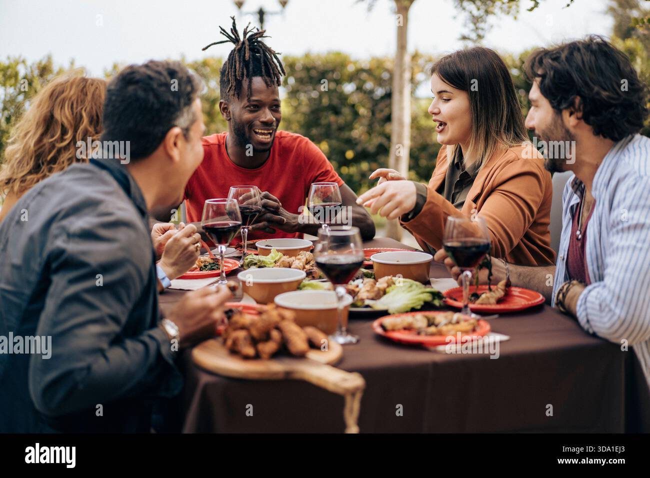Eine Gruppe von Freunden sitzt am Tisch, spricht und genießt Wein bei einem entspannten Abendessen im Freien. Warme Atmosphäre, echte Emotionen, vielfältige Menschen, Essen Stockfoto