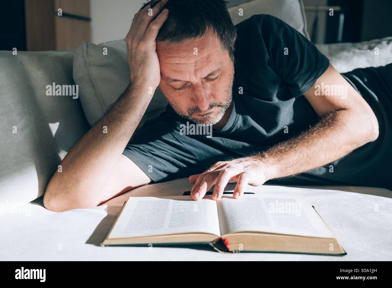 Authentischer Mann mittleren Alters, der auf der Couch liegt, erschöpft und tief in Gedanken aussieht, während er ein offenes Buch liest, Hand auf Stirn. Perfekt zum Überdenken Stockfoto