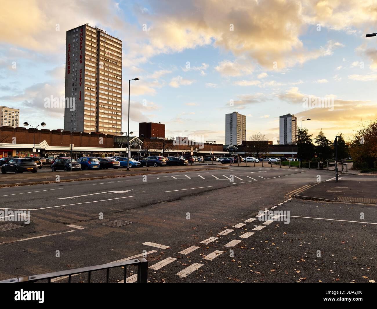 Salford, Vereinigtes Königreich - 8. November 2025: Stadtstraße in Salford mit einem hohen Turm, Geschäften, parkenden Autos und einem hellen Himmel. Stockfoto