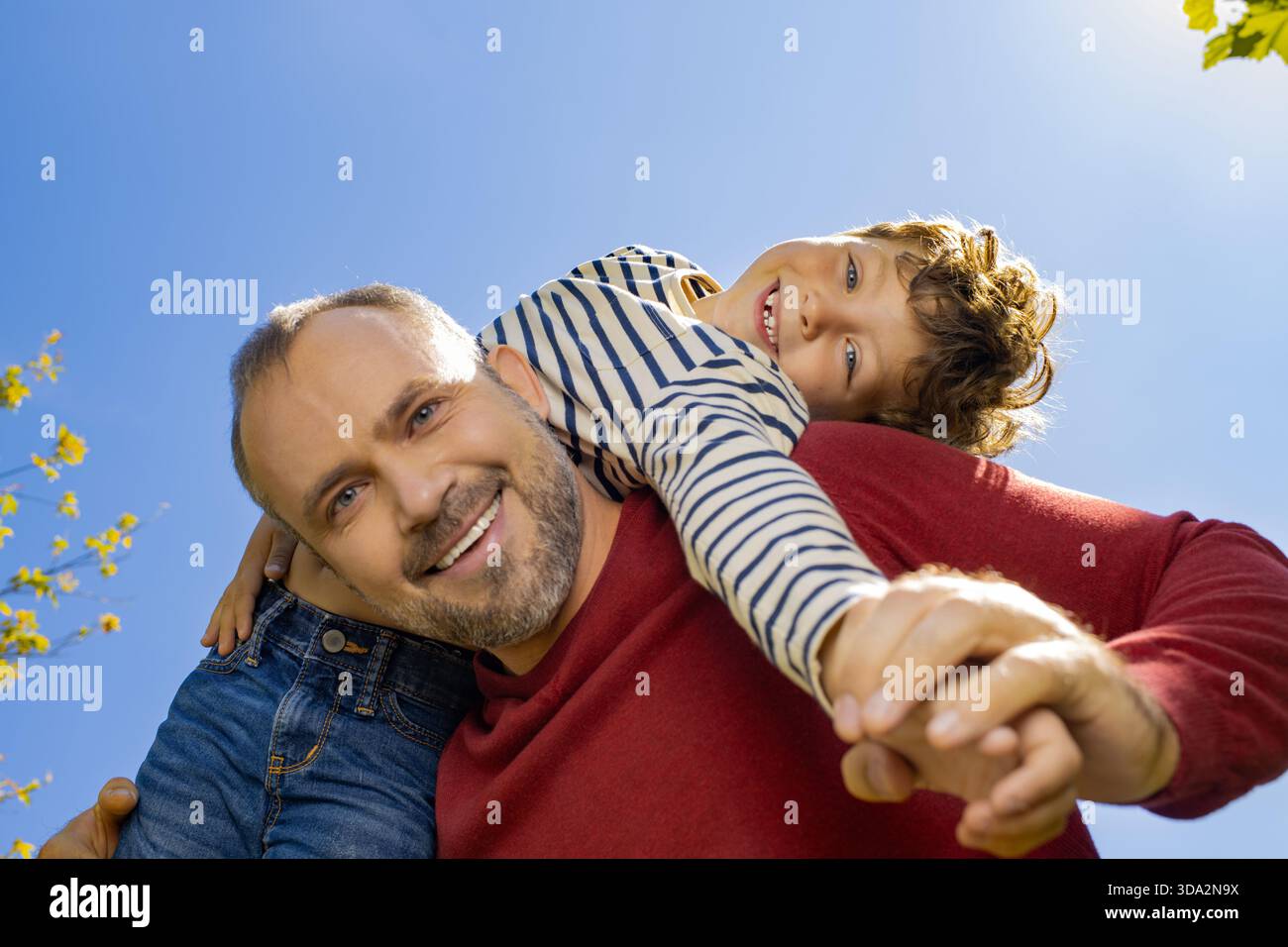 Ein Vater hebt seinen lächelnden Sohn auf die Schultern, umgeben von strahlendem Sonnenschein und Grün. Stockfoto