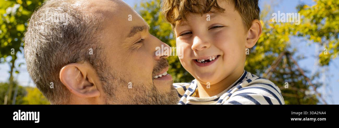 Ein Vater lächelt seinen fröhlichen Sohn an, beide kneifen sich in der Wärme eines sonnigen Tages draußen. Stockfoto
