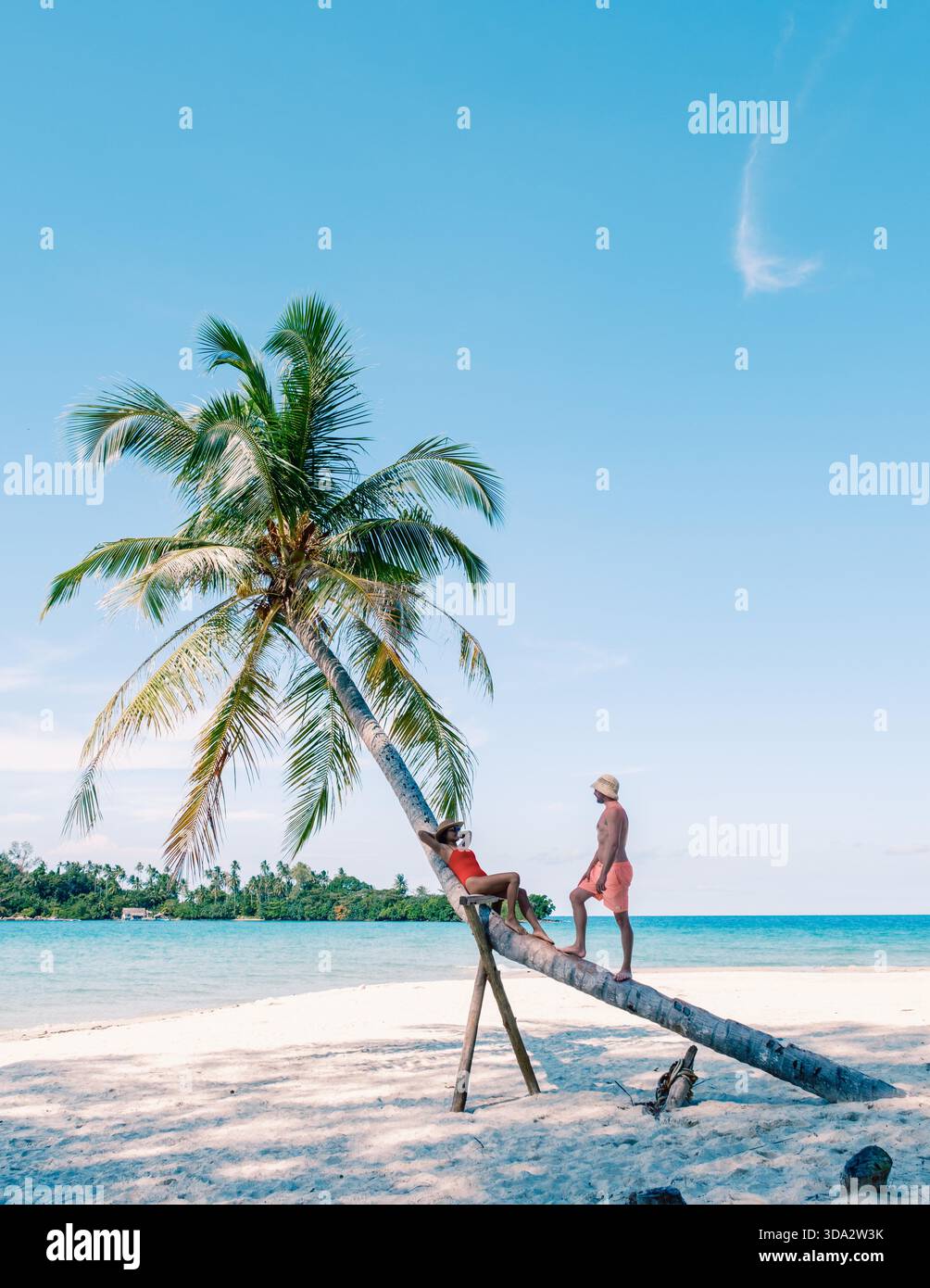 Wunderschöne Strandszene in Koh Kood, Thailand, mit einem ruhigen Moment, in dem sich eine Frau an einer Palme entspannt, während der Mann sich darauf vorbereitet, als Paar beizutreten. || Modell freigegeben Stockfoto