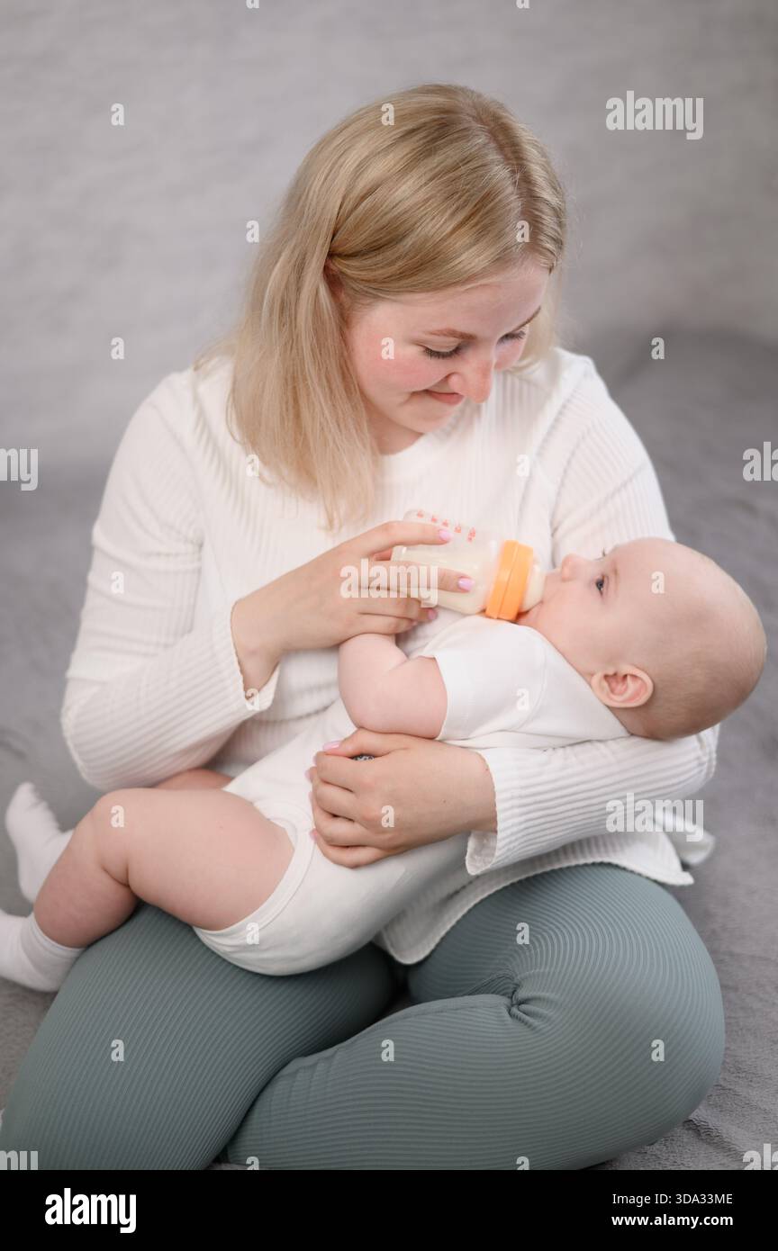 Die Frau sitzt auf dem Bett, hält das Baby in den Armen und ernährt es aus der Flasche. Konzept der Säuglingsfütterung mit der Formel, Alternative zum Stillen. Vertikal Stockfoto