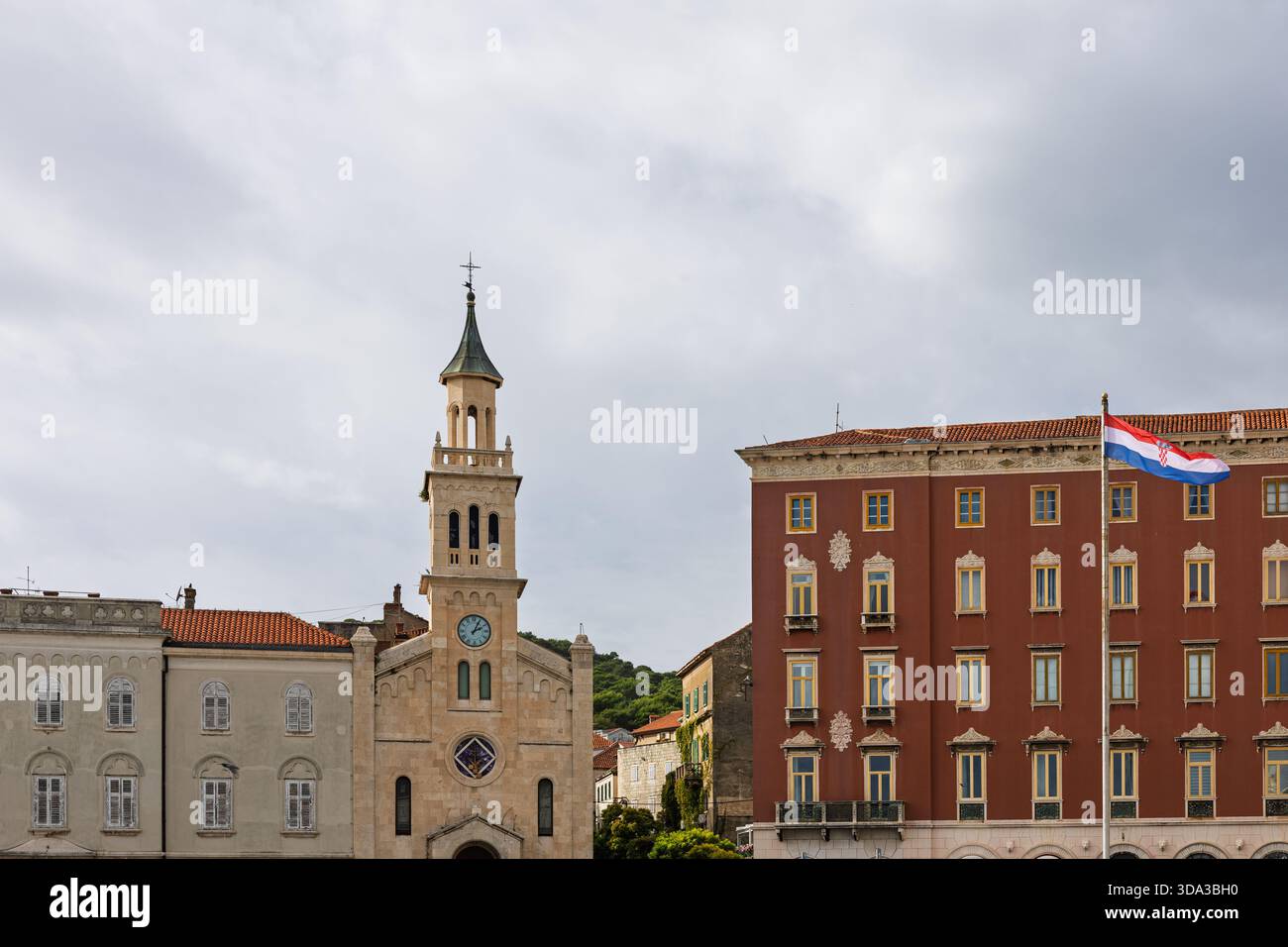 Eine historische europäische Straßenszene mit einer beigefarbenen Steinkirche mit einem hohen Uhrenturm, flankiert von alten Gebäuden Stockfoto