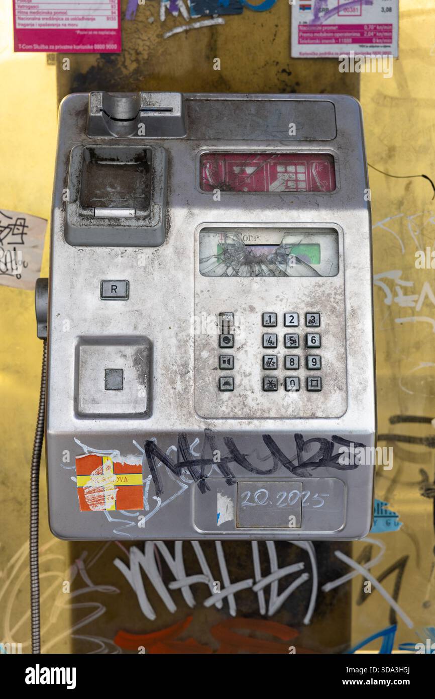 SPLIT, KROATIEN, 10.25.2025: Ein verwittertes Münztelefon sitzt an einer mit Graffiti bedeckten gelben Wand, umgeben von Plakaten und bunten Tags. Das Retro-Gerät Stockfoto