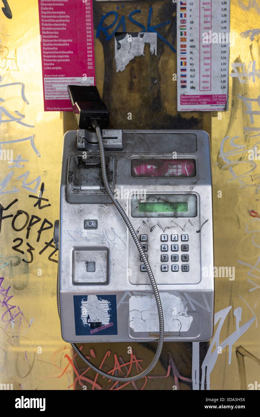 SPLIT, KROATIEN, 10.25.2025: Ein verwittertes Münztelefon sitzt an einer mit Graffiti bedeckten gelben Wand, umgeben von Plakaten und bunten Tags. Das Retro-Gerät Stockfoto