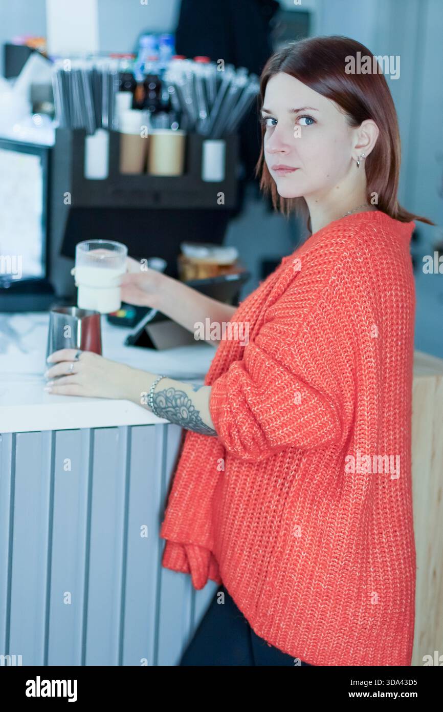 Weibliche Barista in einem pulsierenden orangefarbenen Pullover gießt Milch in den Pitcher an der modernen Kaffeekasse, wobei verschiedene Werkzeuge und Gegenstände im Blick sind. Stockfoto