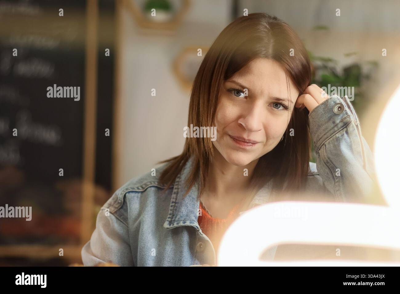 Brünette Frau in lässigem Outfit zeigt ein ruhiges, sanftes Lächeln, während sie das Haar in einem gemütlichen Café mit warmem Umgebungslicht berührt Stockfoto