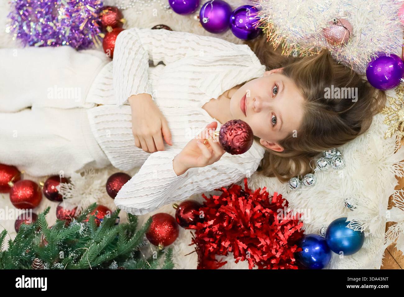 Das Kind trägt weiße Strickwaren und hält glitzernde Verzierungen, während es auf einem Pelzteppich mit Weihnachtsschmuck in der Nähe liegt Stockfoto