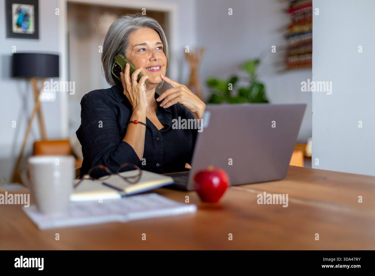 Lächelnde, reife Geschäftsfrau, die auf dem Mobiltelefon spricht und wegblickt, während sie zu Hause aus der Ferne arbeitet Stockfoto