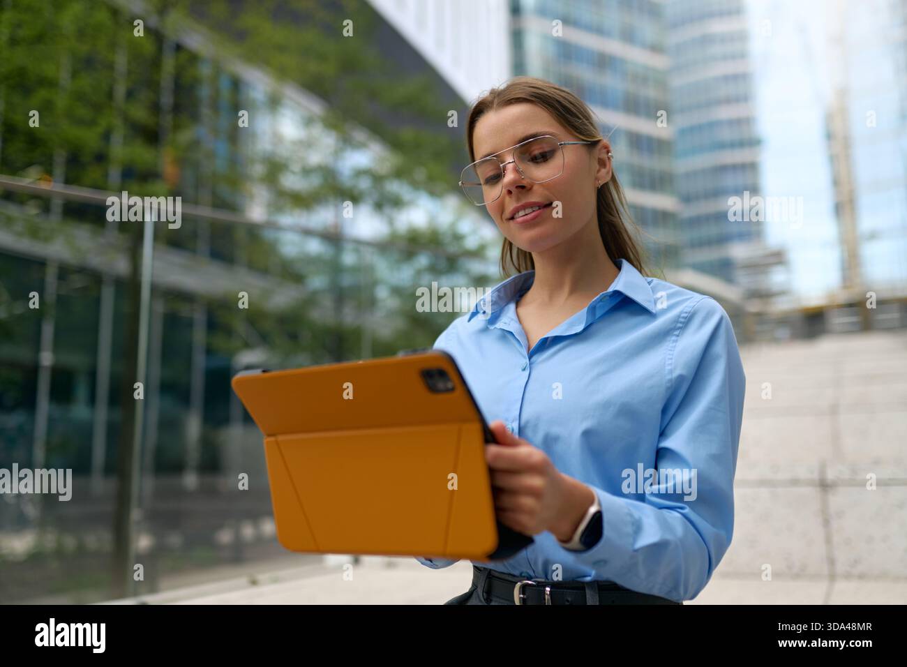 Junge Frau in blauem Hemd und Brille lächelt, während sie draußen an einem Tablet mit orangefarbenem Gehäuse arbeitet, städtische Glasgebäude im Hintergrund. Moderne Busi Stockfoto