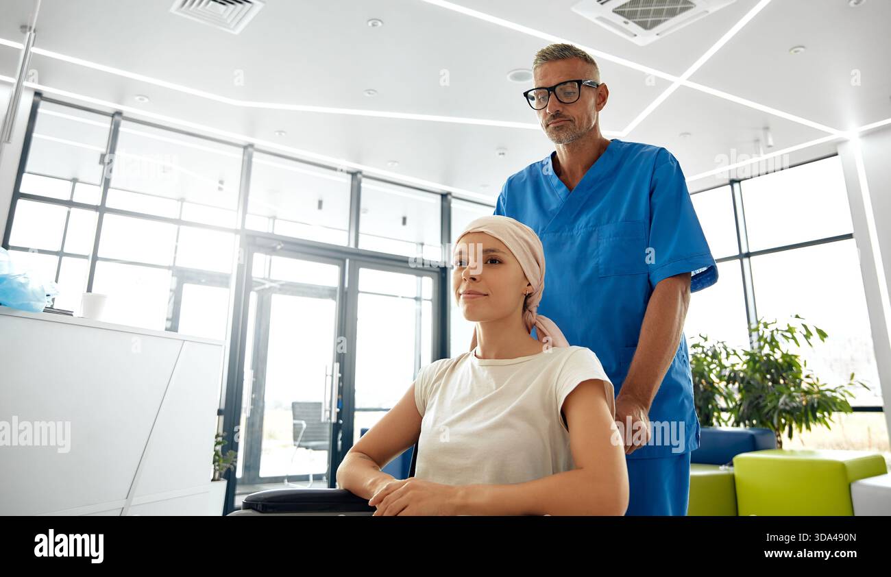 Ein engagiertes medizinisches Fachpersonal, das in blauen Peelings gekleidet ist, hilft mitfühlend einer Patientin, die in einem Rollstuhl an einer modernen medizinischen Einrichtung sitzt Stockfoto