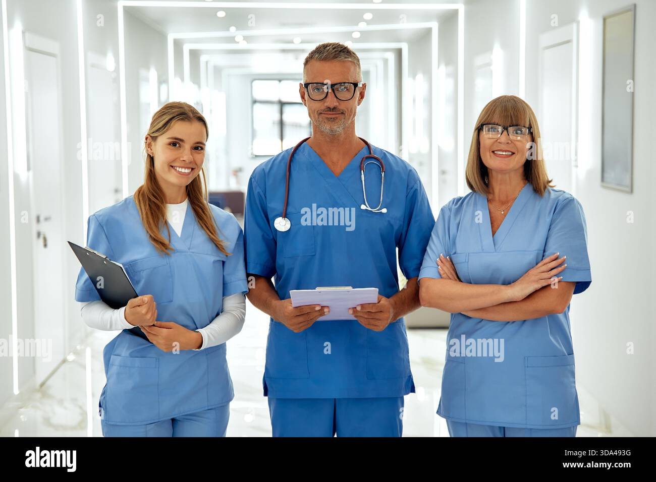 Ein engagiertes Gesundheitsteam von drei lächelnden Personen in hellblauen Peelings ist bereit, Patienten in einem einladenden Krankenhausflur zu helfen, strahlend Stockfoto