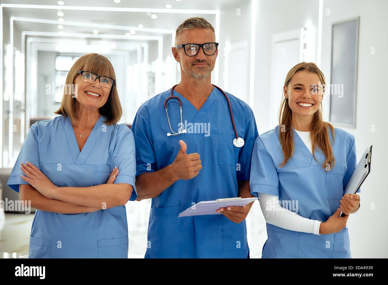 Ein engagiertes Gesundheitsteam von drei lächelnden Personen in hellblauen Peelings ist bereit, Patienten in einem einladenden Krankenhausflur zu helfen, strahlend Stockfoto