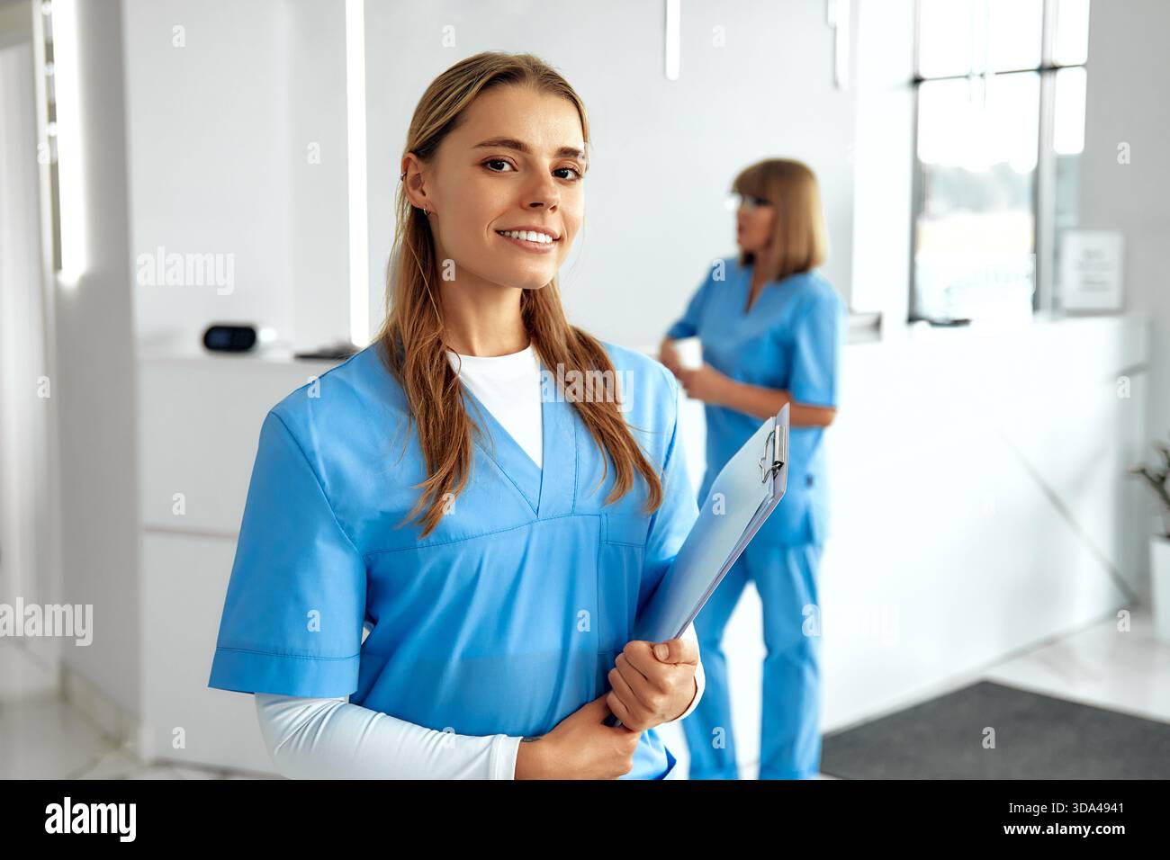 In einem hellen Gesundheitsumfeld lächelt eine selbstbewusste Krankenschwester in blauer Farbe, während sie ein Klemmbrett hält, und ein anderer Mitarbeiter arbeitet, um sicherzustellen, dass die Patienten in der Hand sind Stockfoto