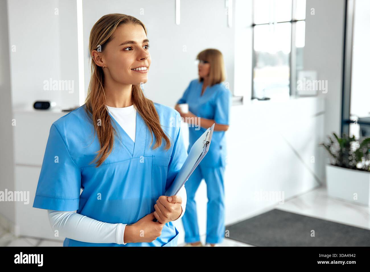 In einem hellen Gesundheitsumfeld lächelt eine selbstbewusste Krankenschwester in blauer Farbe, während sie ein Klemmbrett hält, und ein anderer Mitarbeiter arbeitet, um sicherzustellen, dass die Patienten in der Hand sind Stockfoto