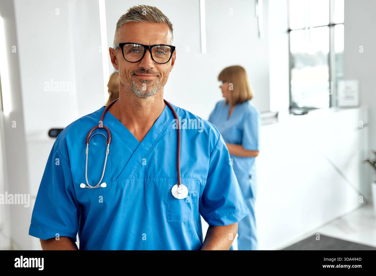 Ein sehr selbstbewusster männlicher Arzt, gekleidet mit blauen Peelings und Brille, steht in einem modernen Gesundheitsumfeld, voll vorbereitet zu helfen Stockfoto
