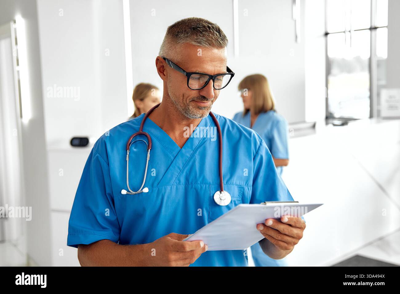 Ein sehr selbstbewusster männlicher Arzt, gekleidet mit blauen Peelings und Brille, steht in einem modernen Gesundheitsumfeld, voll vorbereitet zu helfen Stockfoto