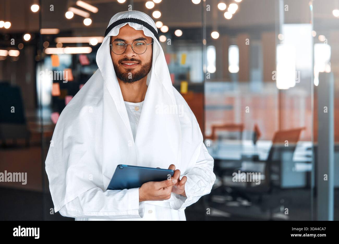 Junger Geschäftsmann aus dem Nahen Osten in traditioneller emiratischer Kleidung mit einem Tablet im Büro. Geschäftsteams arbeiten und Brainstorming in UA Stockfoto