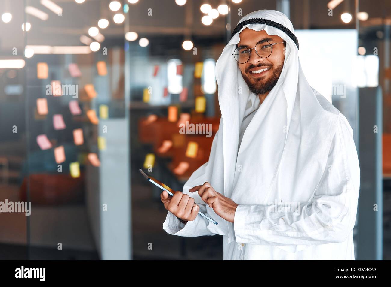 Junger Geschäftsmann aus dem Nahen Osten in traditioneller emiratischer Kleidung mit einem Tablet im Büro. Geschäftsteams arbeiten und Brainstorming in UA Stockfoto