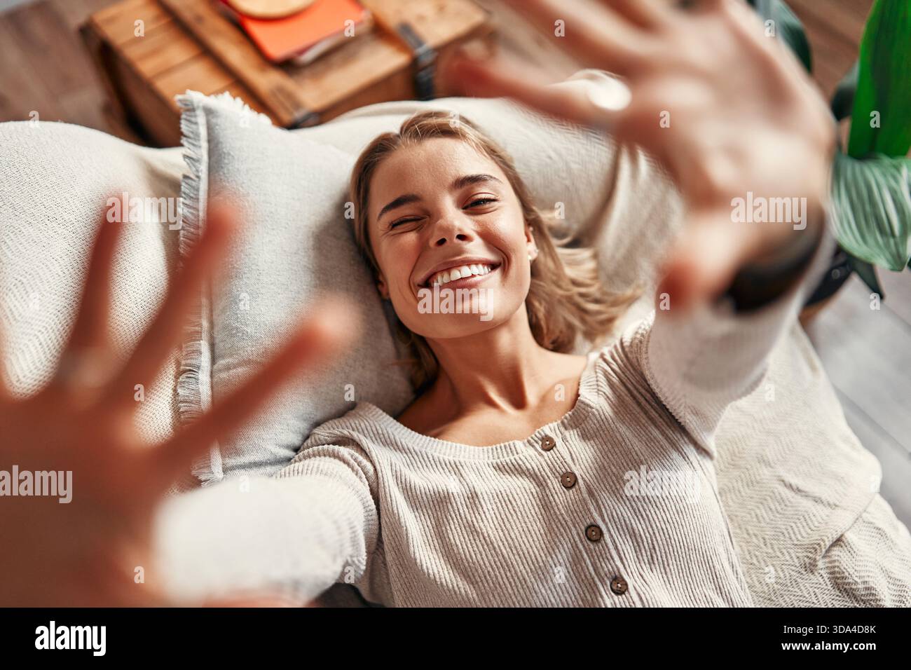 Junge Frau, die auf einem Sofa in einem gemütlichen Wohnzimmer zu Hause liegt und sich an einem freien Tag entspannt, lächelt und zur Kamera greift. Stockfoto