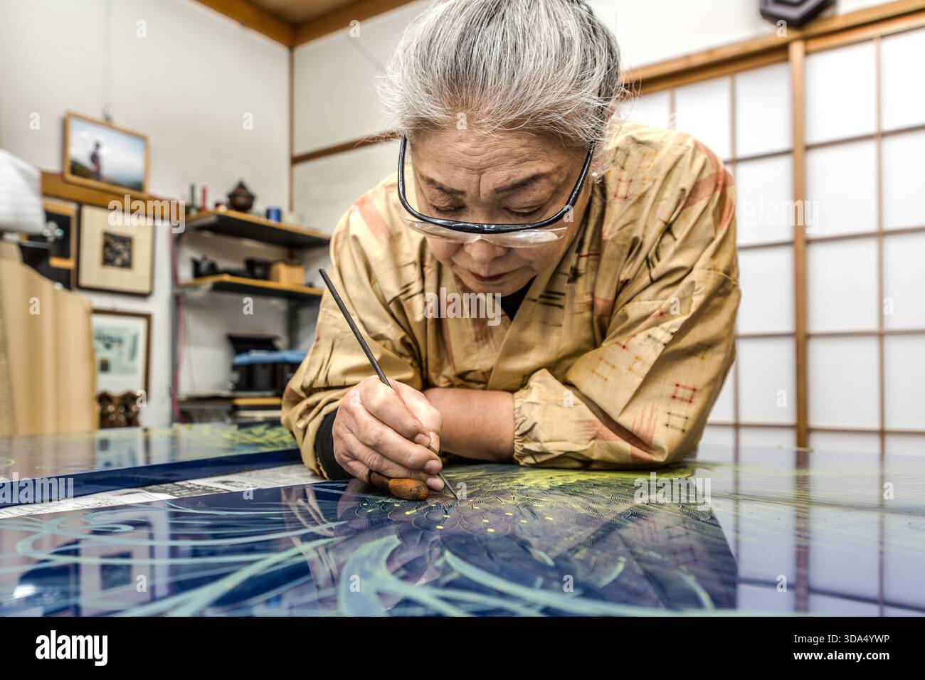 Eine ältere Lackkünstlerin mit Brille arbeitet intensiv an einer großformatigen blauen Lackplatte in ihrem Atelier, die mit einem feinen Messer florale und ornamentale Motive in sie einschneidet. Shiojiri, Präfektur Nagano, Japan Stockfoto