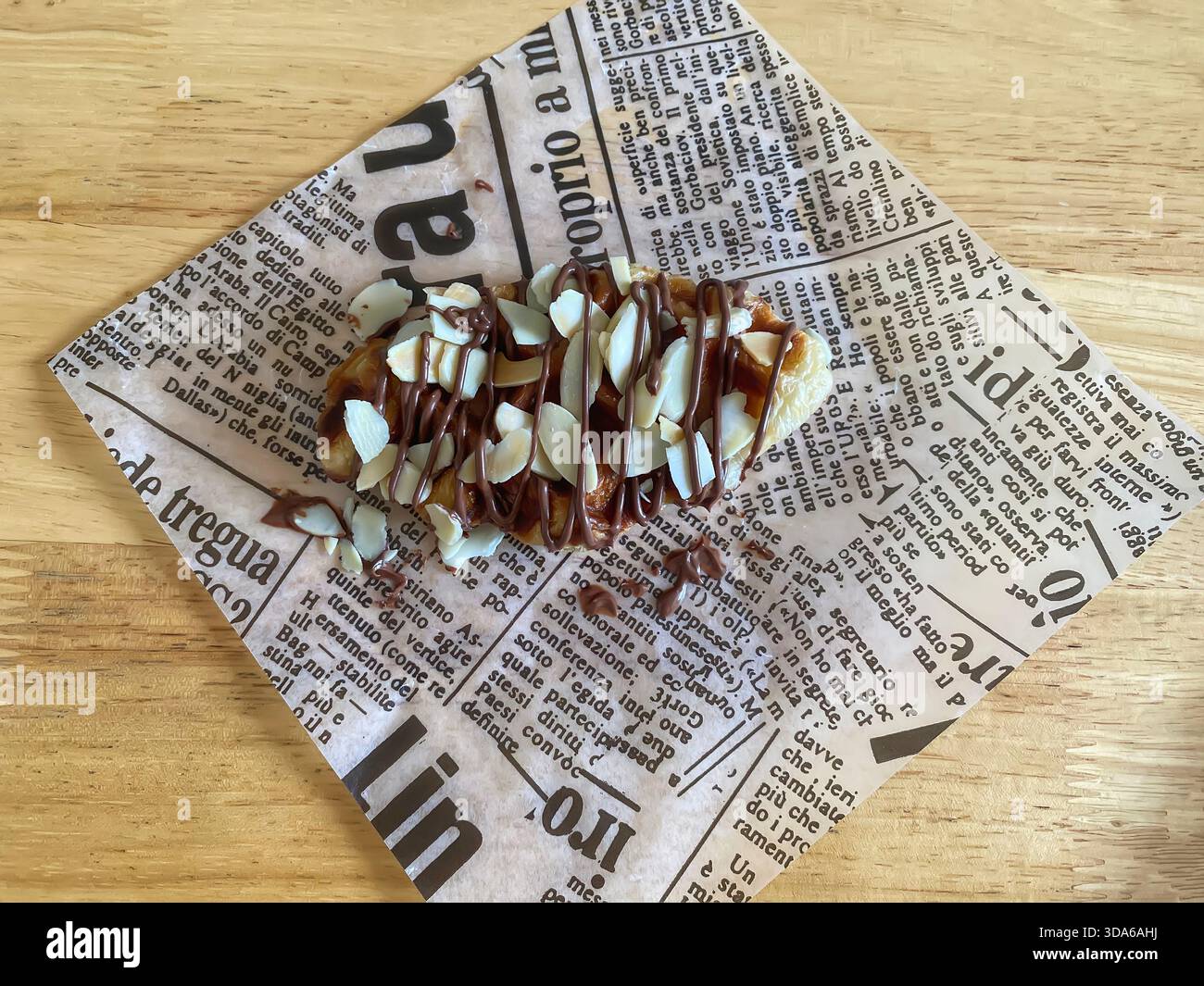 Ein knuspriges Croissant-Waffel oder -Croffle mit Schokoladengläser und Mandelscheiben, serviert auf einem bedruckten Papierbogen auf einem Holztisch. Stockfoto