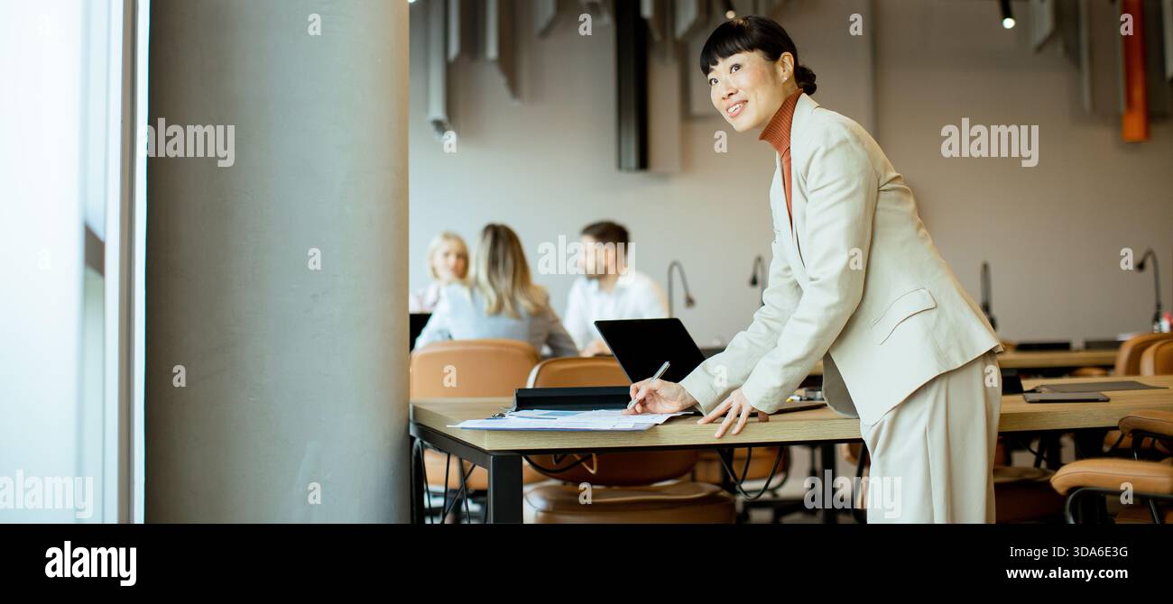In einem lebhaften Büro schreibt eine Frau in einem Anzug an einen Tisch, während Kollegen sich unterhalten. Die Atmosphäre ist produktiv und konzentriert. Stockfoto
