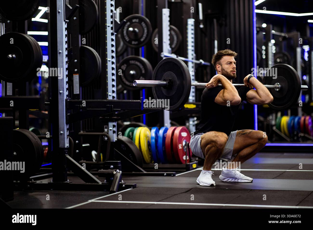 Ein Mann konzentriert sich intensiv, während er eine Langhantelhocke in einem gut ausgestatteten Fitnessstudio macht. Die kräftigen Gewichte auf den Racks verleihen dem Hintergrund Farbe, Showcasin Stockfoto