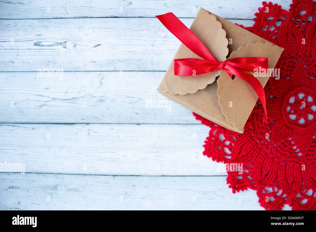 Geschenkbox mit rotem Band auf einem hellroten Häkeldeckchen auf einem rustikalen Holzhintergrund. Minimalistisch handgefertigte festliche Komposition, geeignet für Cell Stockfoto