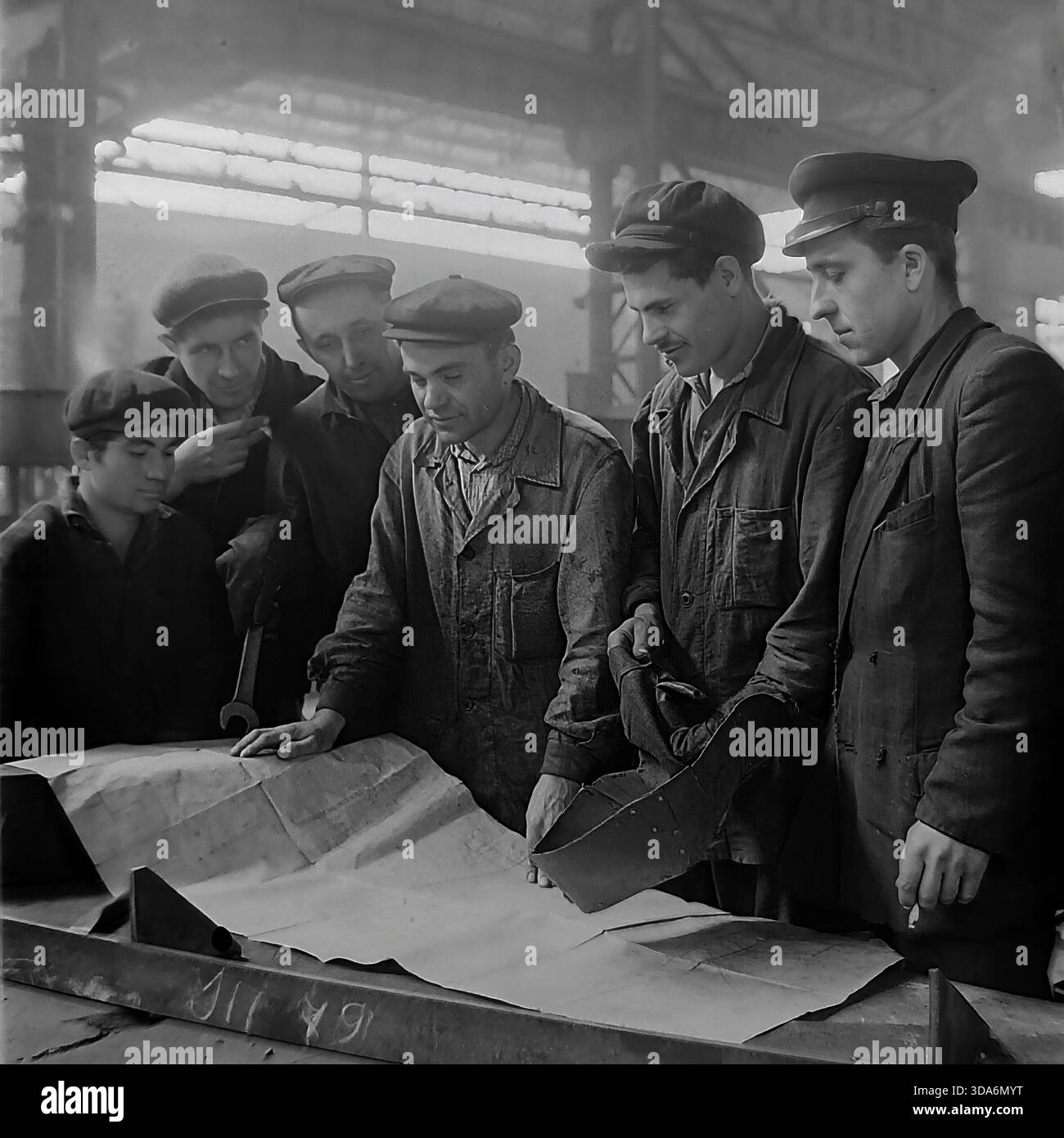 Slawjansk, Ukrainische SSR, UdSSR - 1970er Jahre: Eine Brigade sowjetischer Fabrikarbeiter und Schweißer diskutiert einen technischen Plan im Stroymash-Werk. Eine Gruppe von sechs Männern in Arbeitskleidung und Kappen ist um eine große Zeichnung auf einer Werkbank versammelt. Ein Arbeiter hält eine Schweißmaske in der Hand, während andere den technischen Plan für ein Metallbauprojekt analysieren. Dieses Schwarzweiß-Archivfoto fängt den Geist kollektiver Problemlösung und Industrieplanung in der friedlichen Donbass-Region während der Sowjetzeit ein. Stockfoto