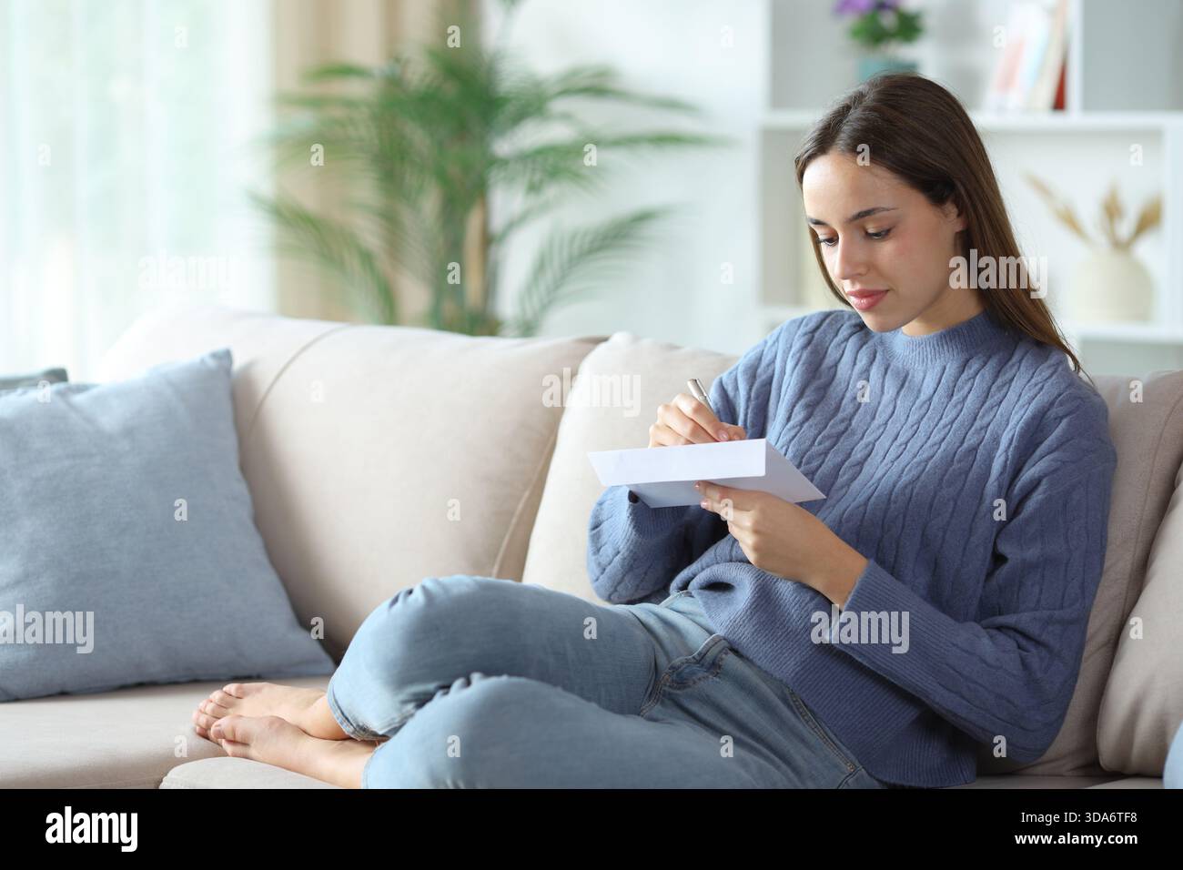 Frau in blauer Schrift Adresse im Umschlag, die zu Hause auf einem Sofa sitzt Stockfoto