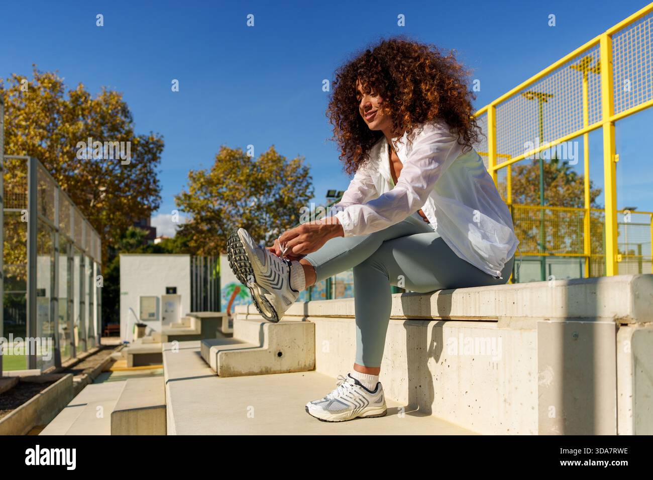Frau mit lockigem Haar, die ihre Schnürsenkel auf Betonstufen bindet, bereitet sich auf eine Outdoor-Fitness oder Padel-Session vor Stockfoto